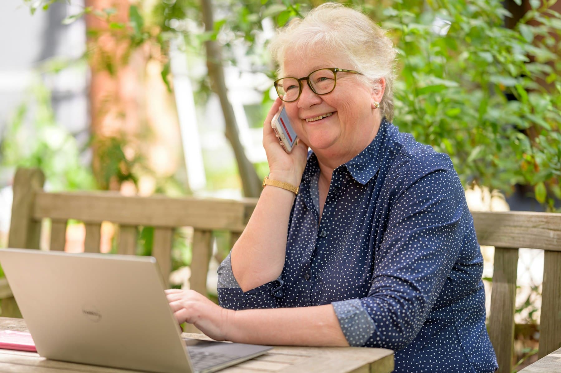 An older woman smiling as she talks on her phone at an outdoor table